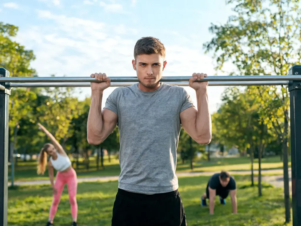 Mężczyzna wykonuje podciągnięcia na drążku w parku, w tle inni ćwiczą, ilustrując trening kalisteniczny dla początkujących.